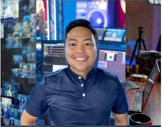 A person smiling in a studio, surrounded by computer monitors displaying digital graphics and various tech equipment.