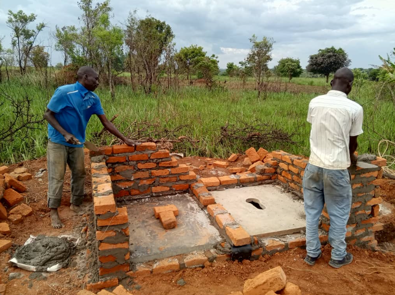 Two people laying bricks to construct an outdoor latrine in a grassy, rural area.