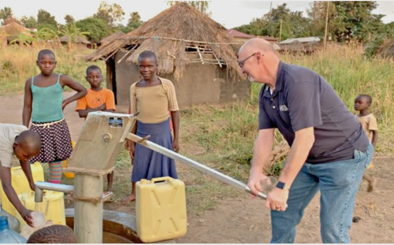 A man is pumping water from a well while children look on.