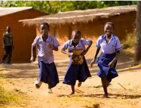 Three students in uniforms running along a dirt path in front of rural buildings.