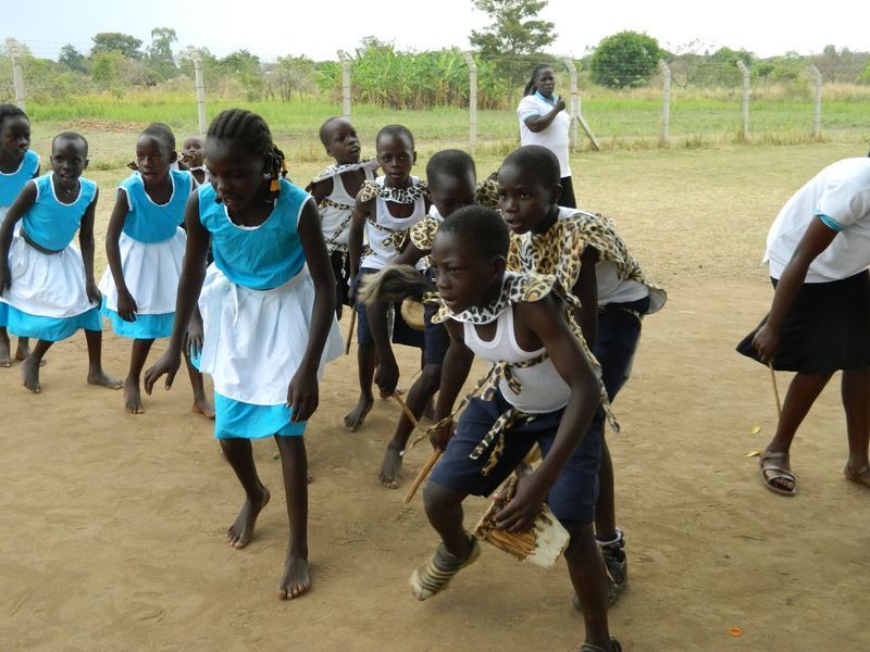 Children dancing outdoors, some wearing blue and white dresses, others with decorative cloths.