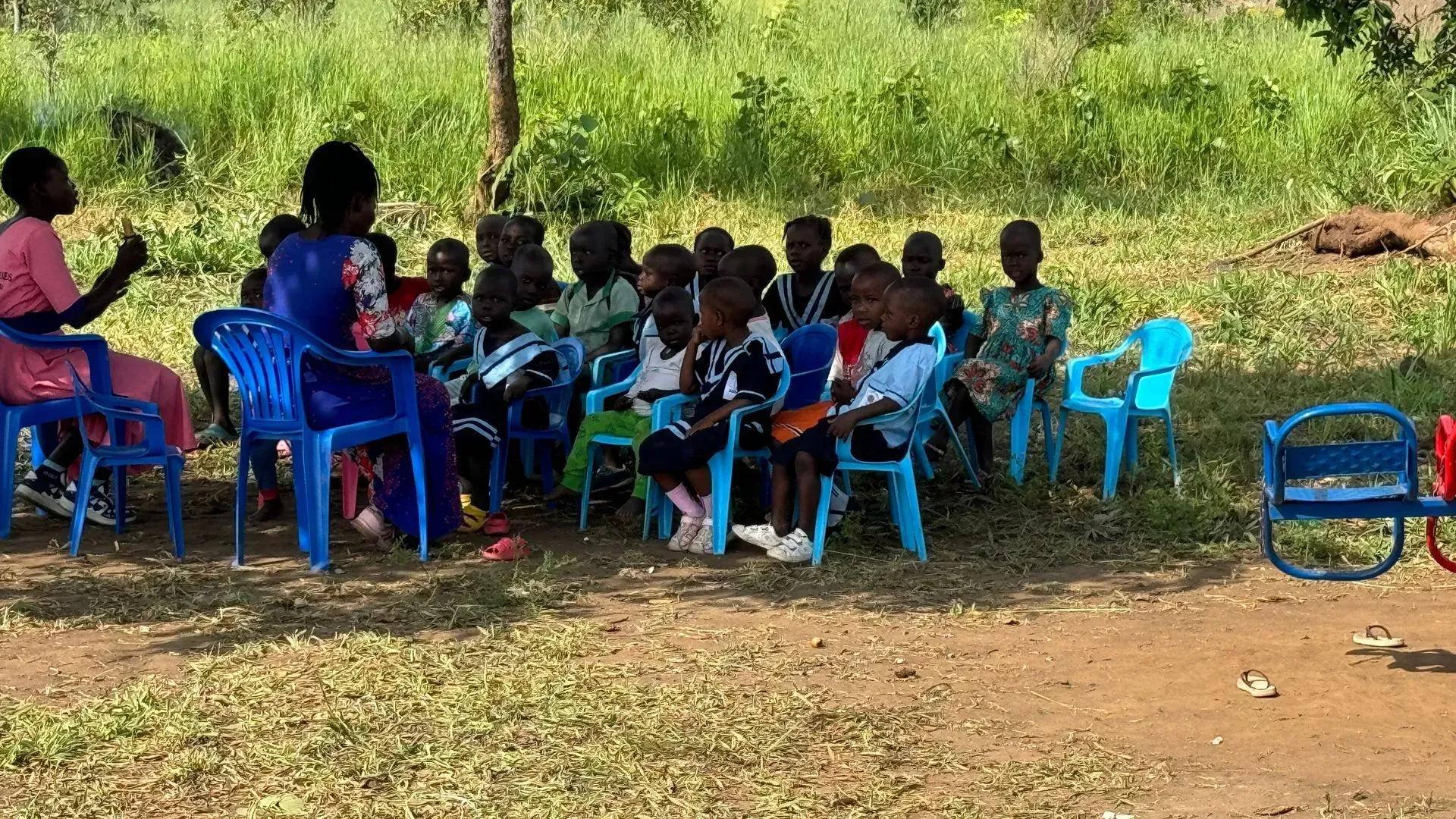 A teacher sits with a group of children in blue plastic chairs during an outdoor class in a grassy, shaded area.
