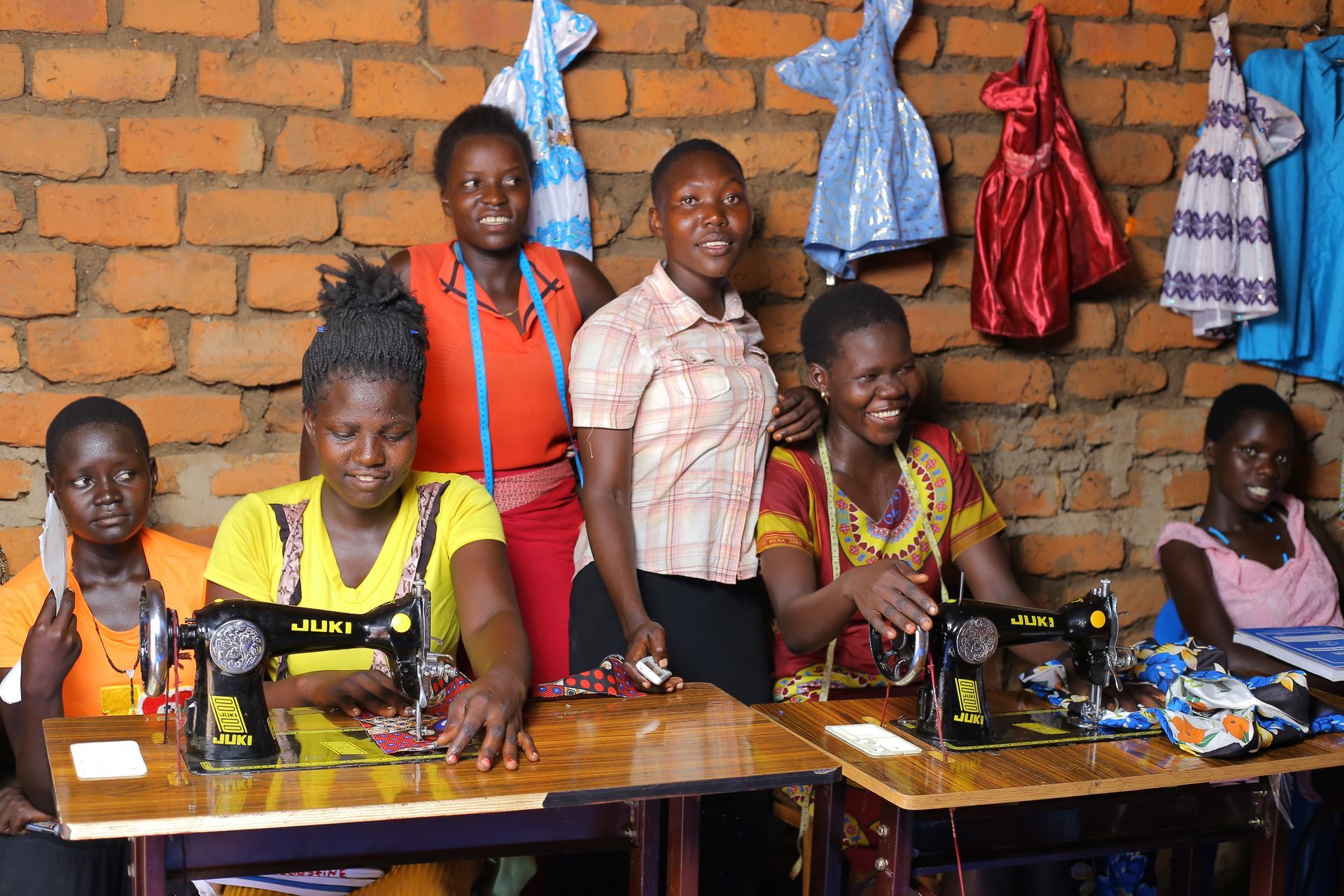 A group of women are sitting at sewing machines in front of a brick wall.