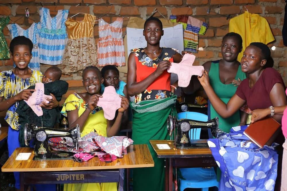 Women in a workshop, holding up handmade cloth sanitary pads, smiling. Sewing machines, colorful clothes.