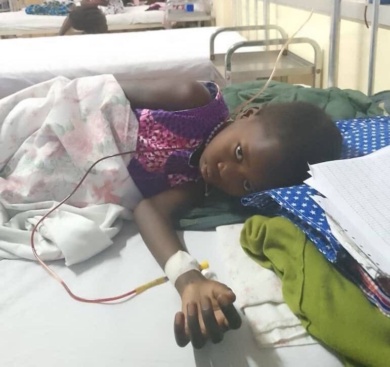 A child in a hospital bed with an IV line in their arm, looking toward the camera.