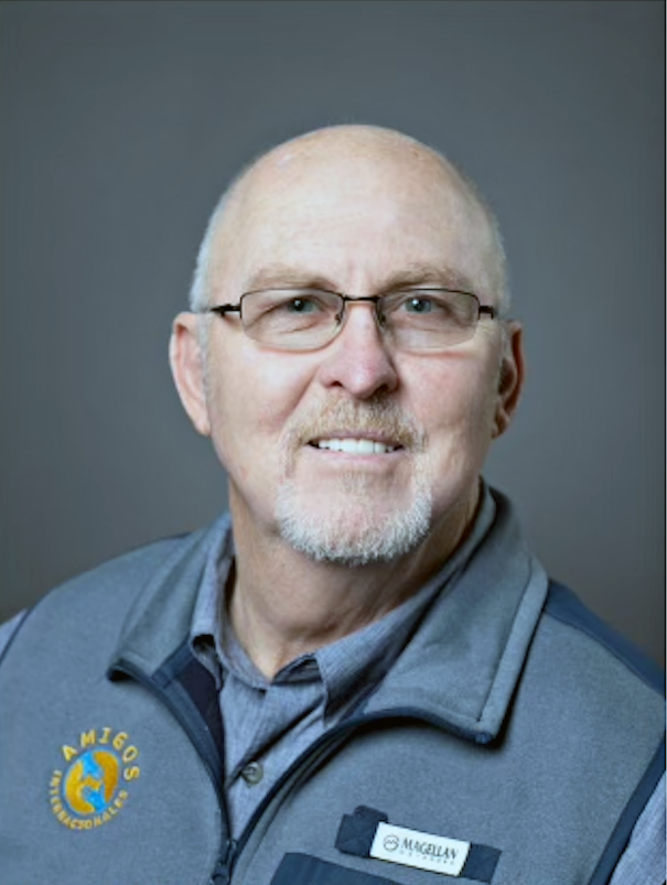 Man with glasses wearing a blue vest, smiling, against a gray backdrop.