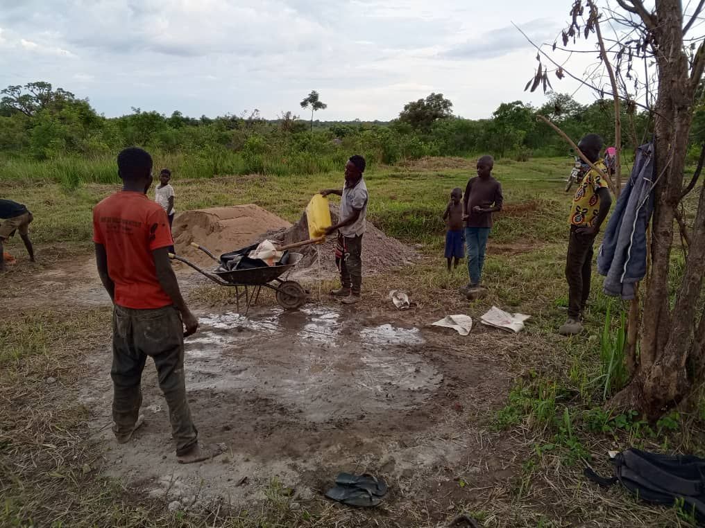 Several people stand in a field with a wheelbarrow near a pile of dirt and a muddy area under an overcast sky.
