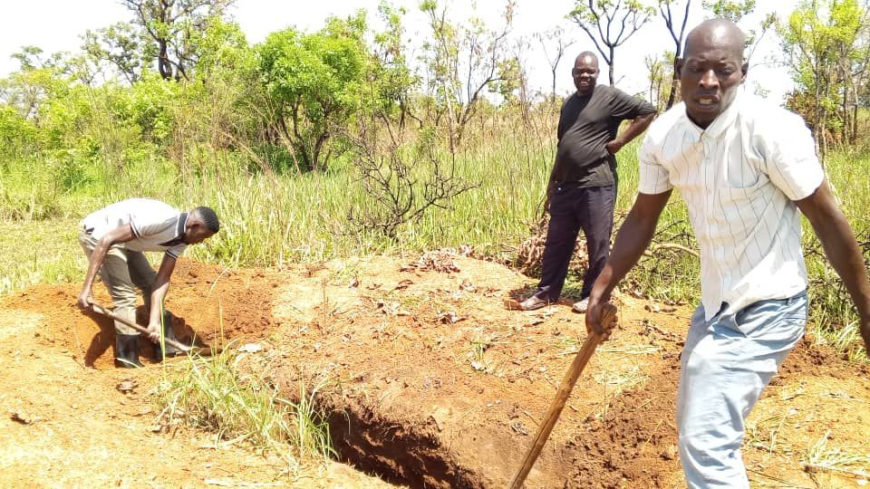 Three people dig a rectangular trench in an outdoor, grassy field with red dirt under a bright, sunny sky.