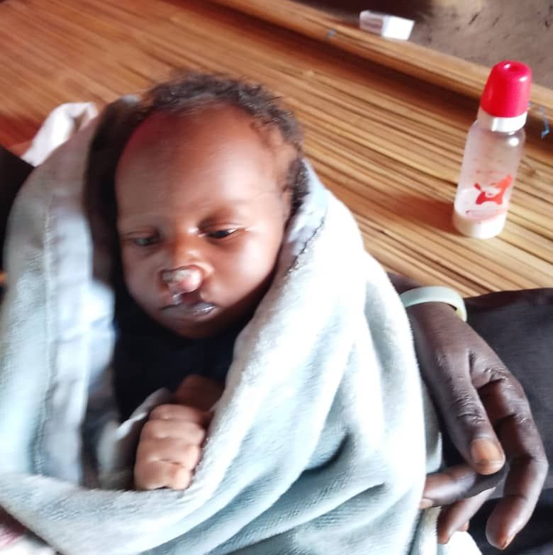Ojok wrapped in a blue blanket, held by someone with dark skin; bottle with red cap nearby.
