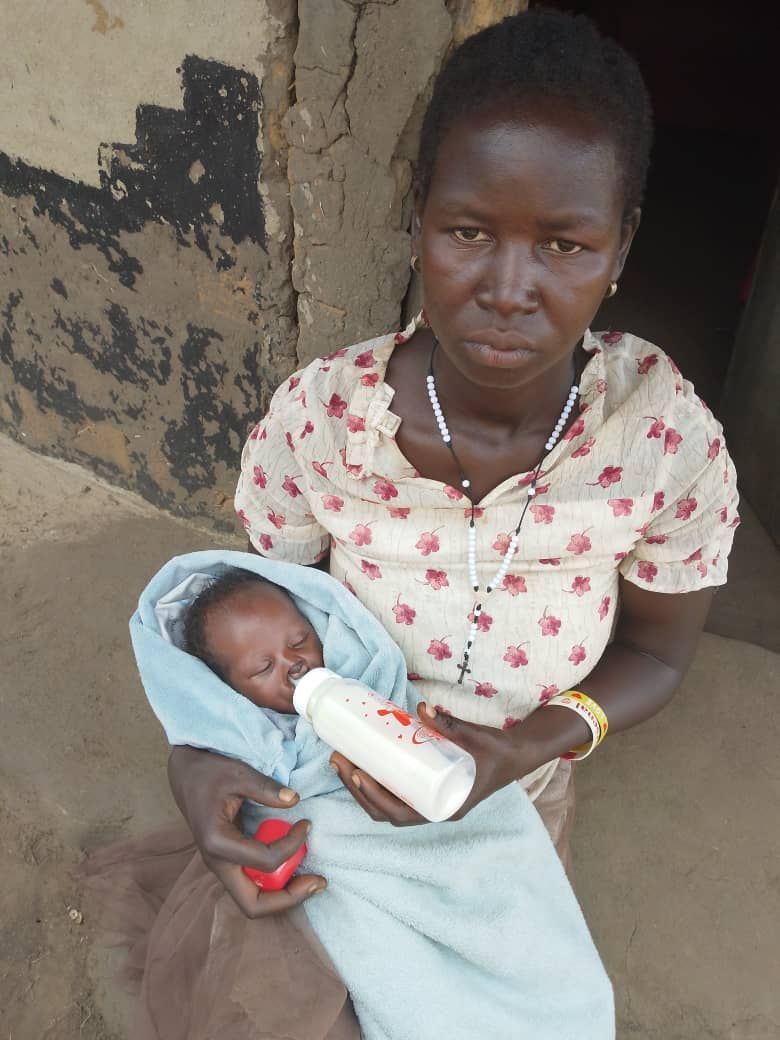 Woman holds a baby, feeding it from a bottle. Both are outside a building.