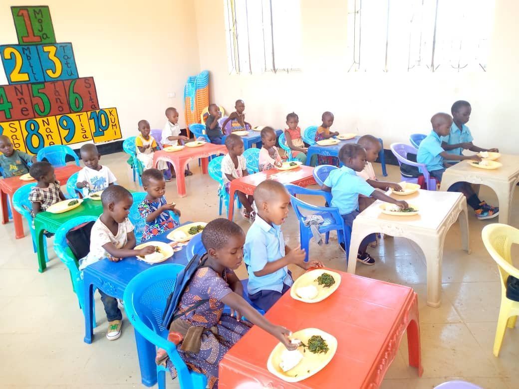 A group of children sits at colorful tables in a classroom, eating from plates during a meal.