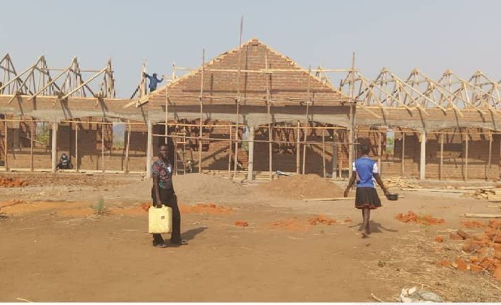 School under construction with workers, children, and building materials in an outdoor setting.