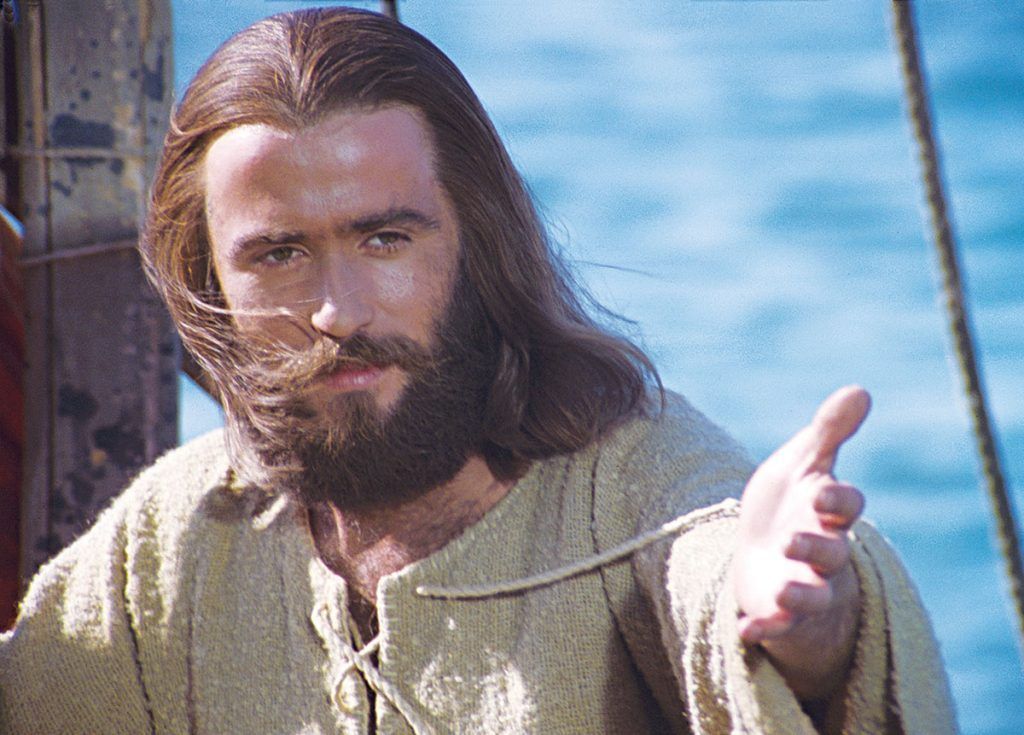 A man with long hair and a beard is standing in front of a body of water.