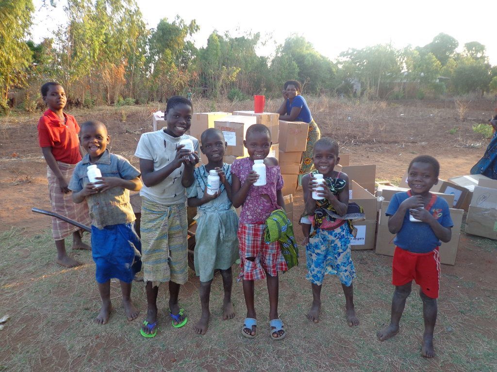 Group of children in an outdoor setting holding small white bottles in front of a stack of cardboard boxes.