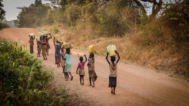 Children walking on a dirt road, carrying water containers on their heads; dry, sunny landscape.