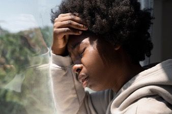 A woman is sitting in front of a window with her hand on her forehead.