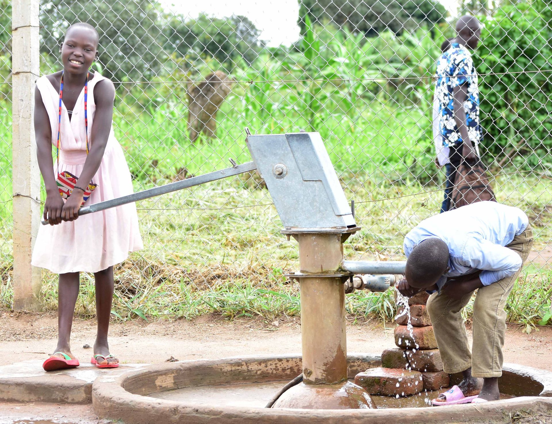 A person pumps a metal hand-operated water well while another person drinks from the water flowing from the spout.