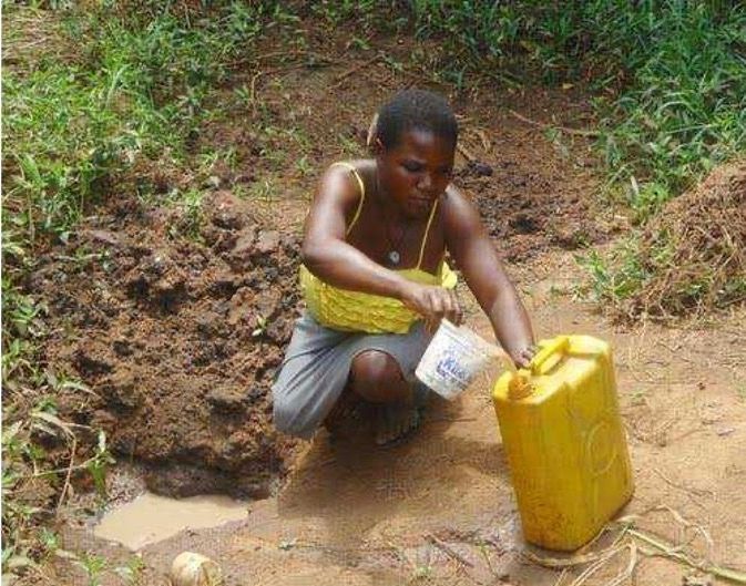 A person crouches outdoors, using a small container to scoop water from a muddy puddle into a yellow plastic jug.