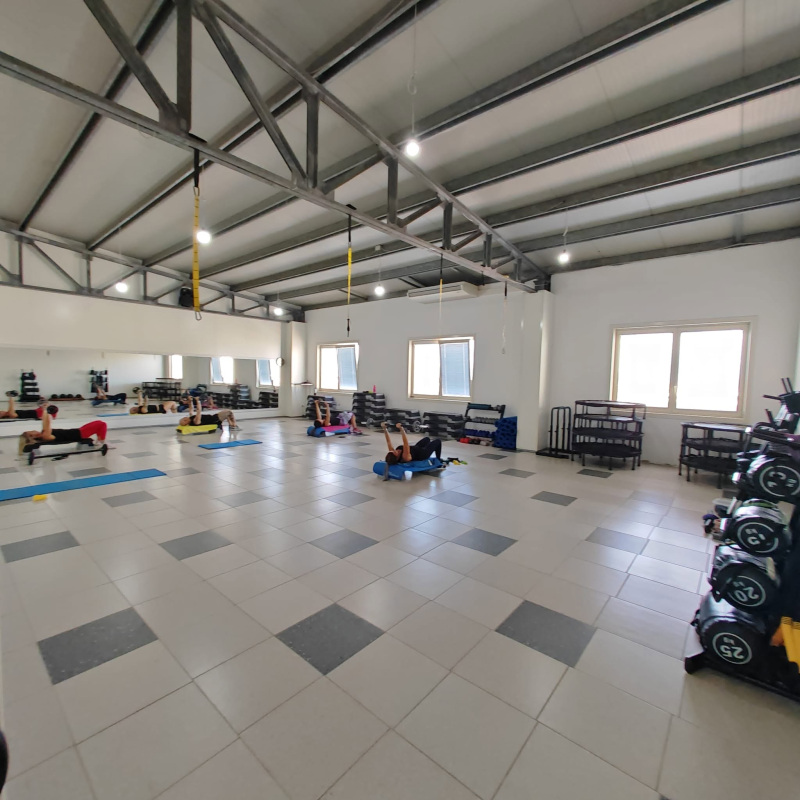Ampia palestra con pavimento piastrellato, rastrelliere per pesi, panche e attrezzature per esercizi sotto un alto tetto in metallo.