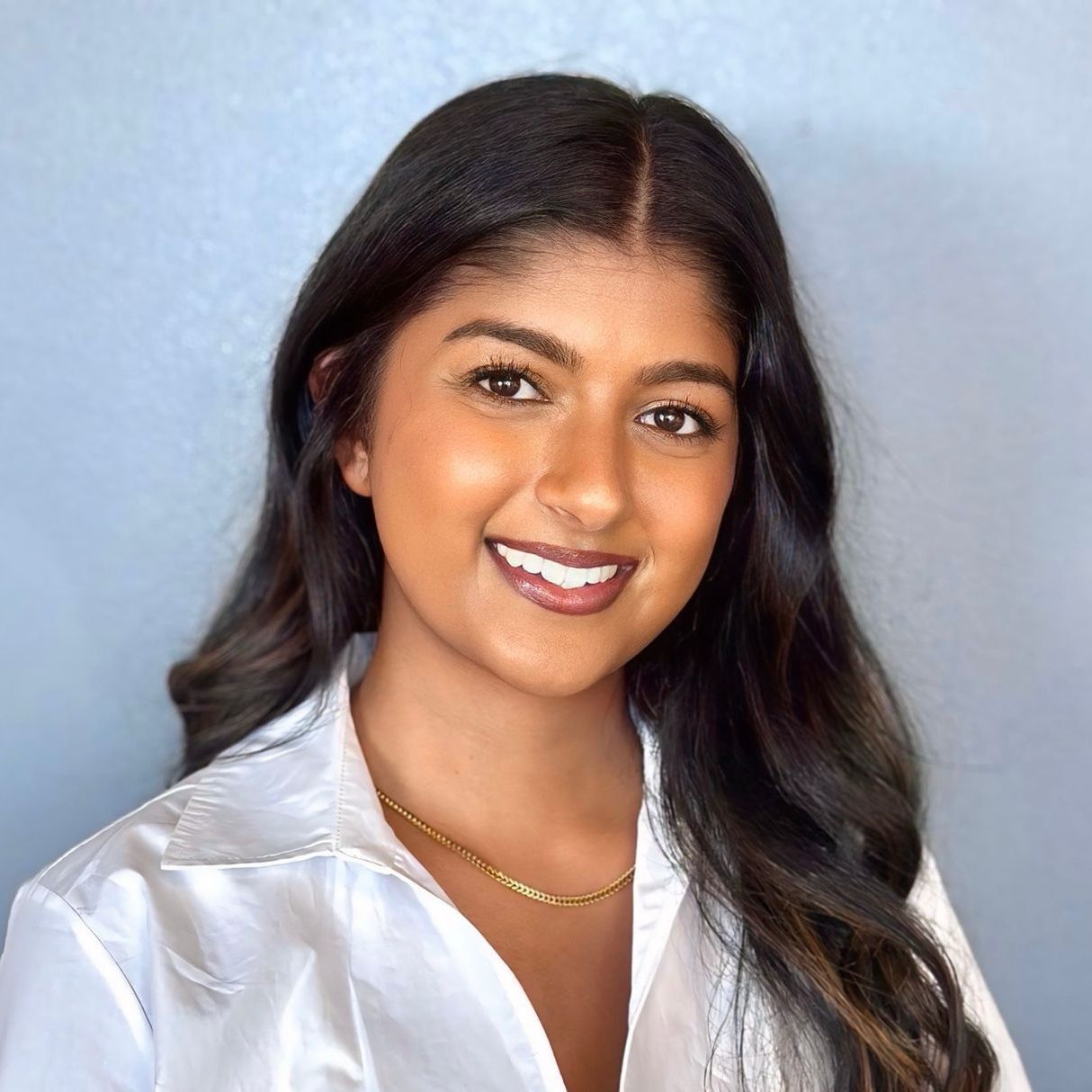 Woman with long dark hair and a white button-down shirt, smiling. She wears a gold necklace against a light blue background.