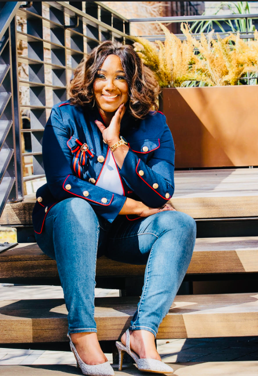 Woman sitting on steps, wearing denim jacket and jeans, smiling, hand on her cheek.