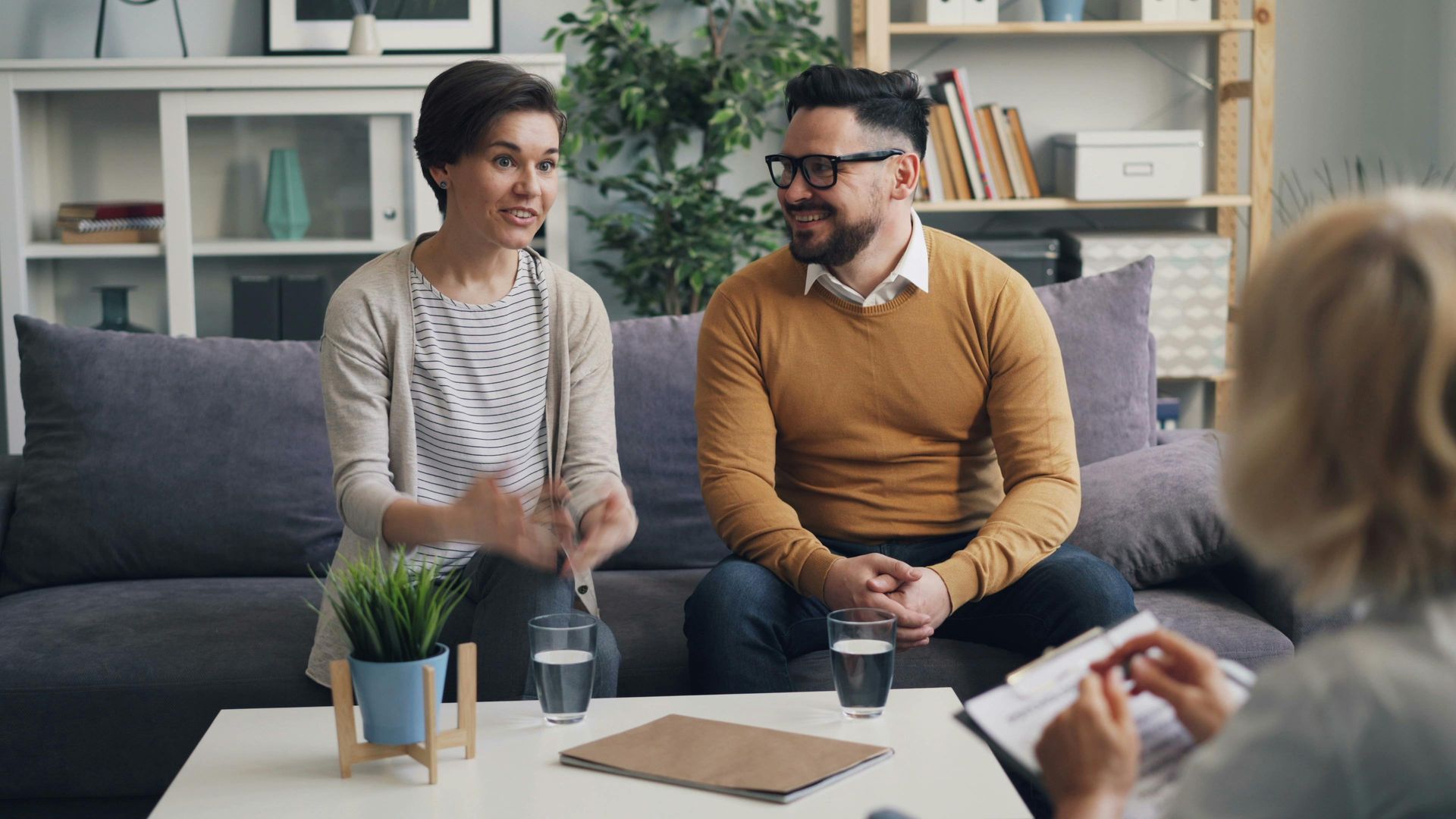 Couple in therapy session, seated on a couch, talking with a therapist in a living room.