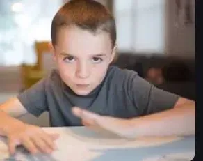 Boy leaning on a table, staring intensely with hands raised. Gray shirt, blurred background.