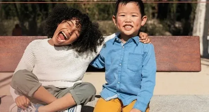 Two children sitting on a bench, one with curly hair laughing, the other with arm around the other child, smiling.