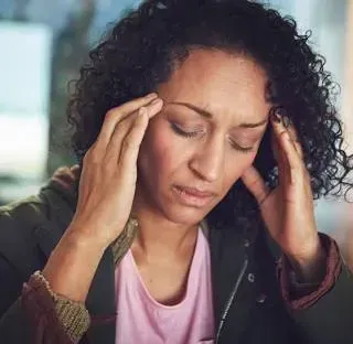 Woman with curly hair, eyes closed, hands on temples, appears to have a headache. Indoors.