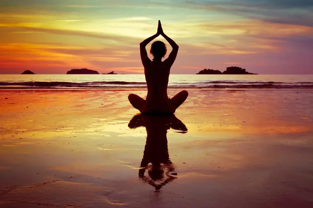 Silhouette of a person in a yoga pose on a beach, hands above head, reflected in wet sand at sunset.