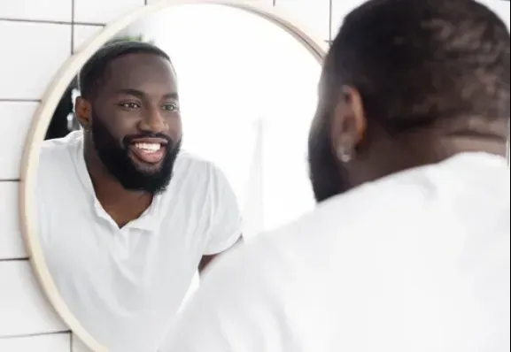 Man smiling at himself in a round mirror, wearing a white shirt in a bathroom.