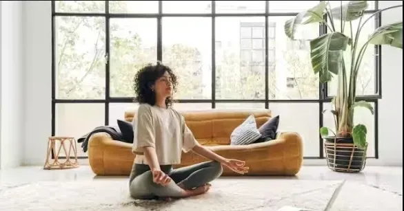 Woman meditating in lotus position on rug in front of a window, with couch and plant visible.