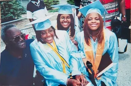 Family at a graduation ceremony, posing for a photo. People in light blue gowns and caps, smiling.