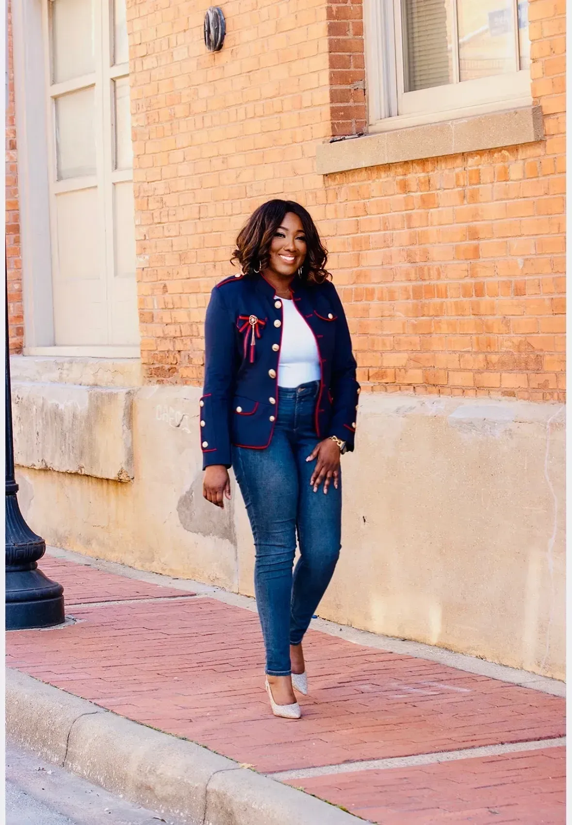 Woman in navy jacket and jeans stands on a brick sidewalk next to a brick building.