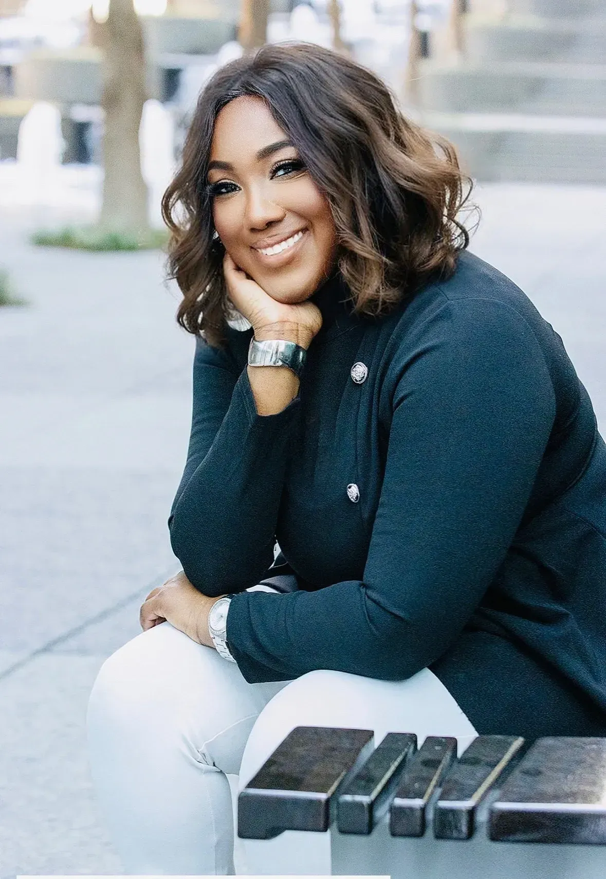 Woman sitting on a bench, smiling. She wears a dark top, white pants, and a silver watch. Outdoors.