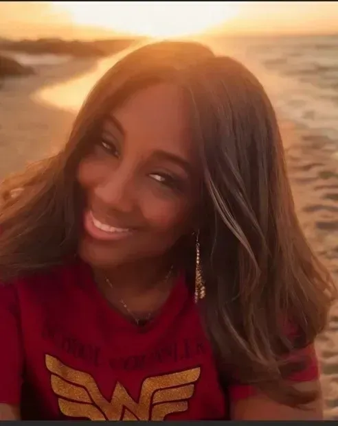 Woman smiling at the beach wearing a red Wonder Woman shirt; sunset in background.