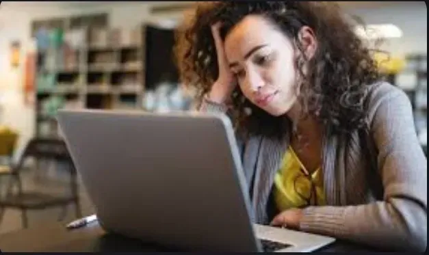 Woman looking at laptop, hand on head, in a library.