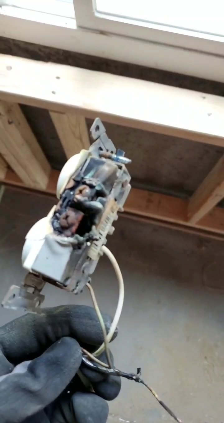 Gloved hand holding a damaged electrical outlet with exposed wires under a ceiling beam