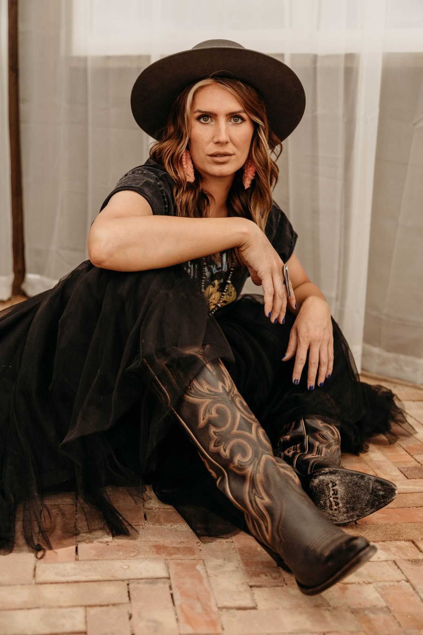 Woman in a black dress and hat sitting on a brick floor by sheer curtains, looking at the camera.