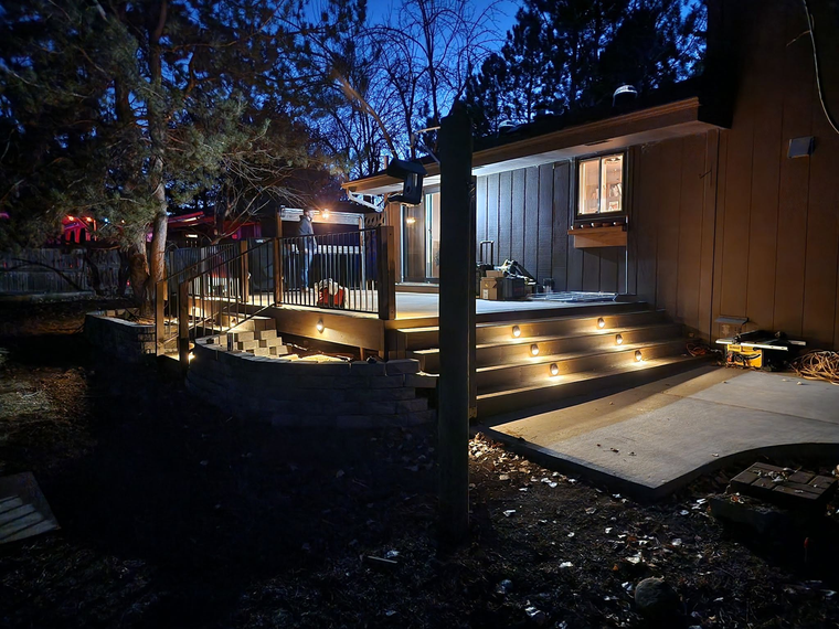 Lit wooden house porch with steps and warm lights at night, beside a concrete path and trees.