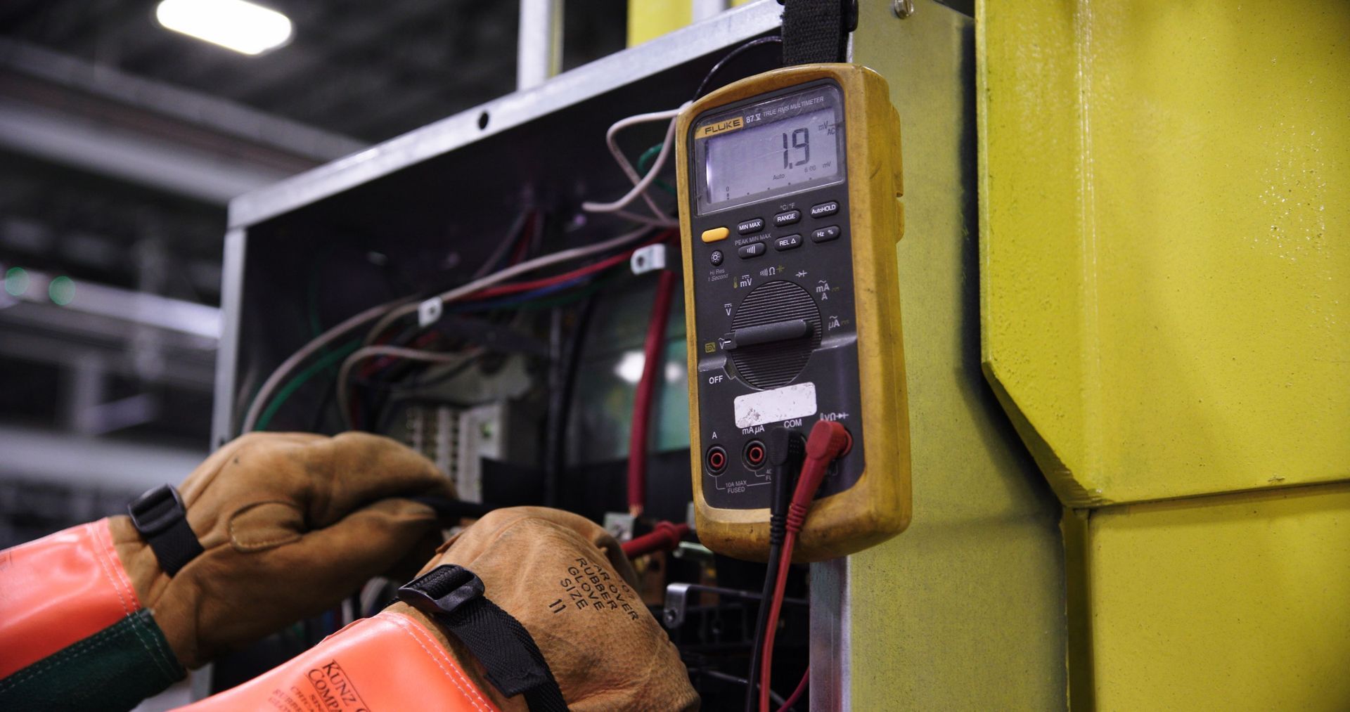 Gloved hands using a multimeter to test wires inside an electrical panel cabinet