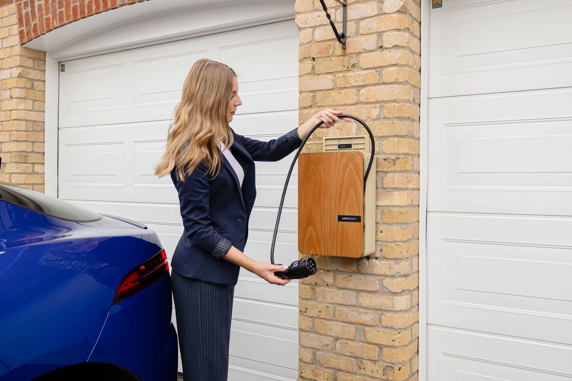 Woman hanging a wooden mailbox on a brick wall beside white garage doors and a blue car