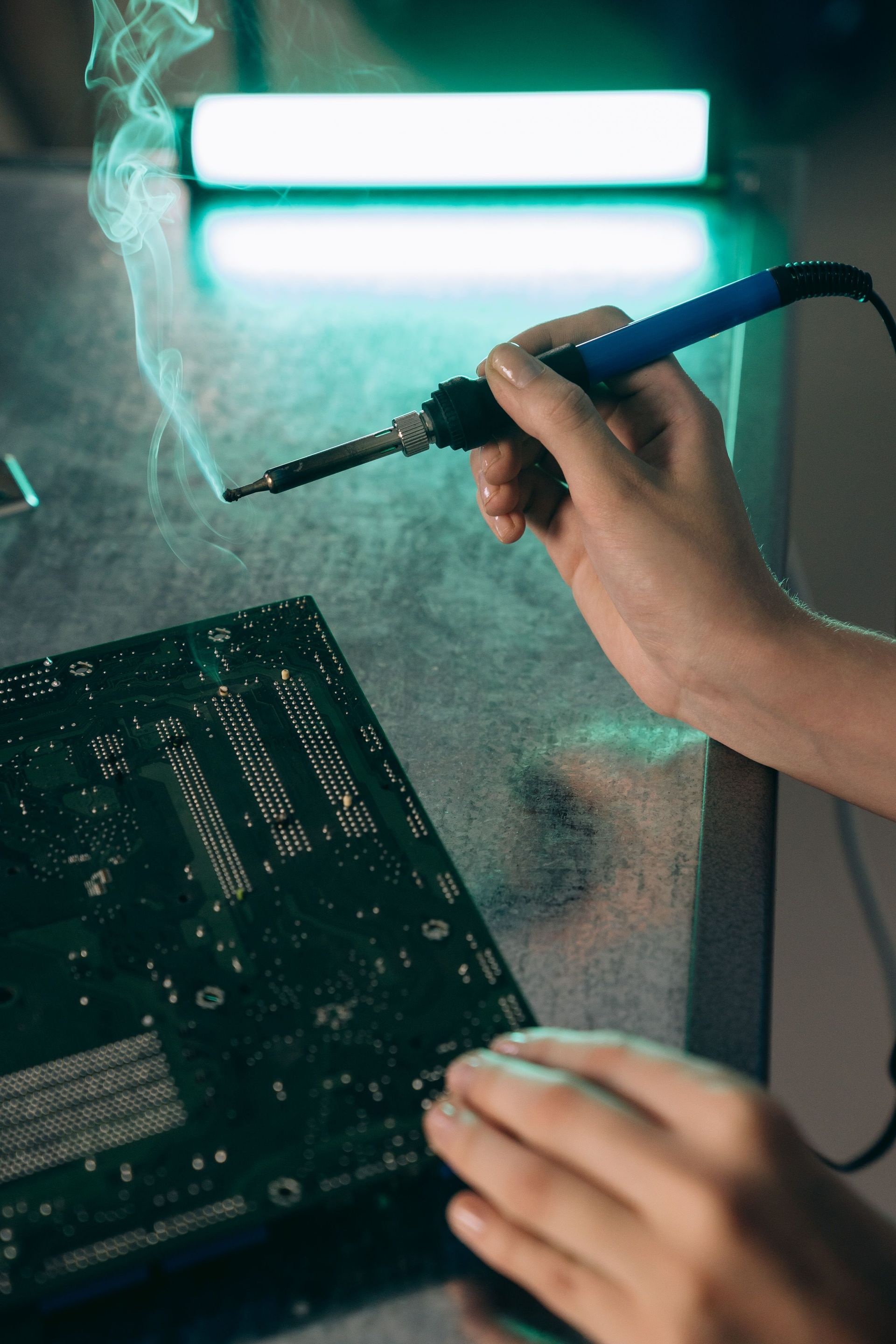 Hands soldering a circuit board with a blue soldering iron in a smoky workshop under bright light