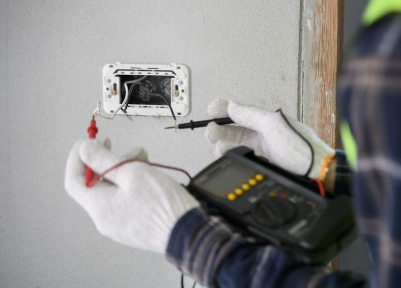 Electrician testing wires in a wall outlet with a multimeter, wearing white gloves.