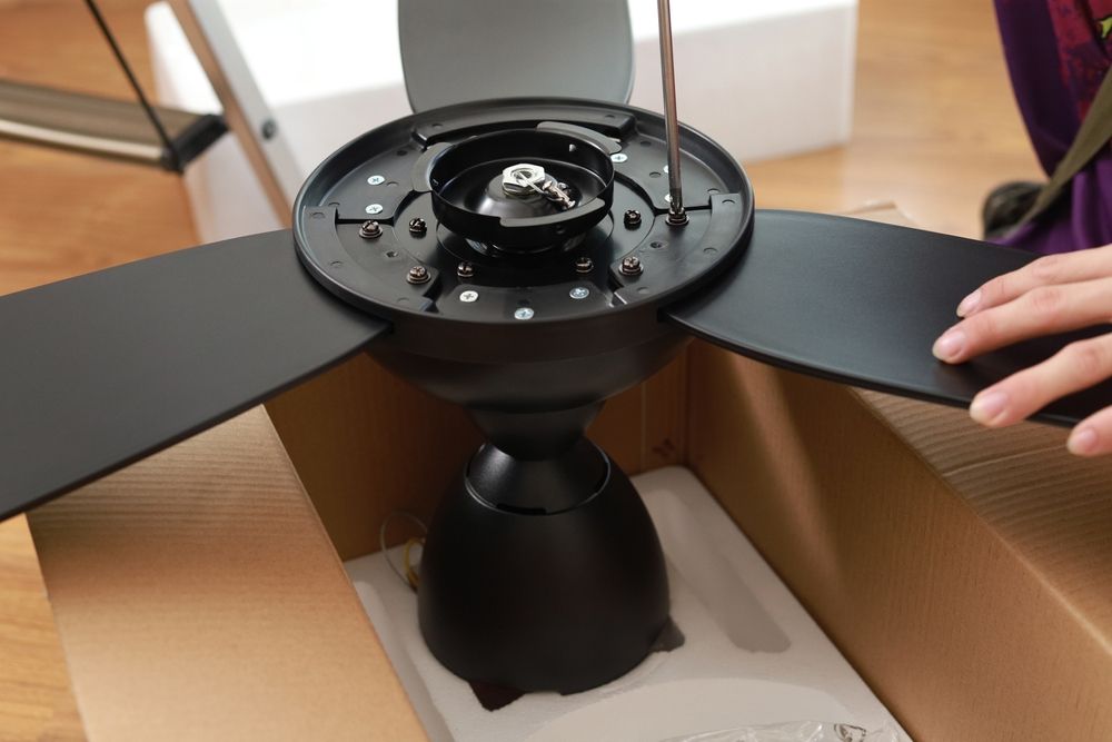 Black ceiling fan with three blades in a cardboard box, with a hand holding one blade.