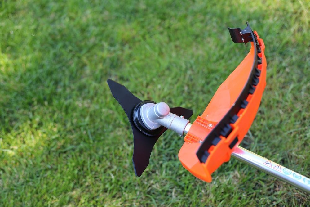 String Trimmer with Black Blade and Orange Guard, Lying on Green Grass — Young's Lawn Care In Aitkenvale, QLD