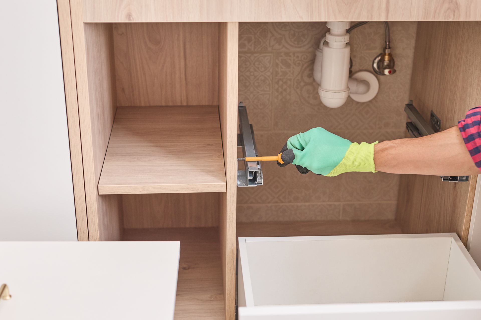 Gloved hand using a screwdriver to install a shelf inside a wooden cabinet under a sink