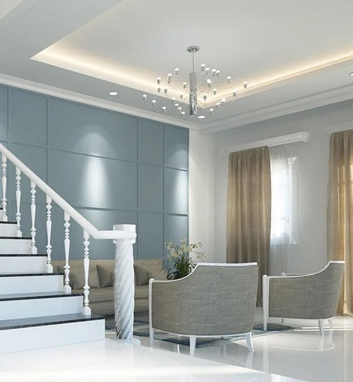 A contemporary living space with a blue paneled wall, a white staircase, two grey armchairs, and a modern chandelier.