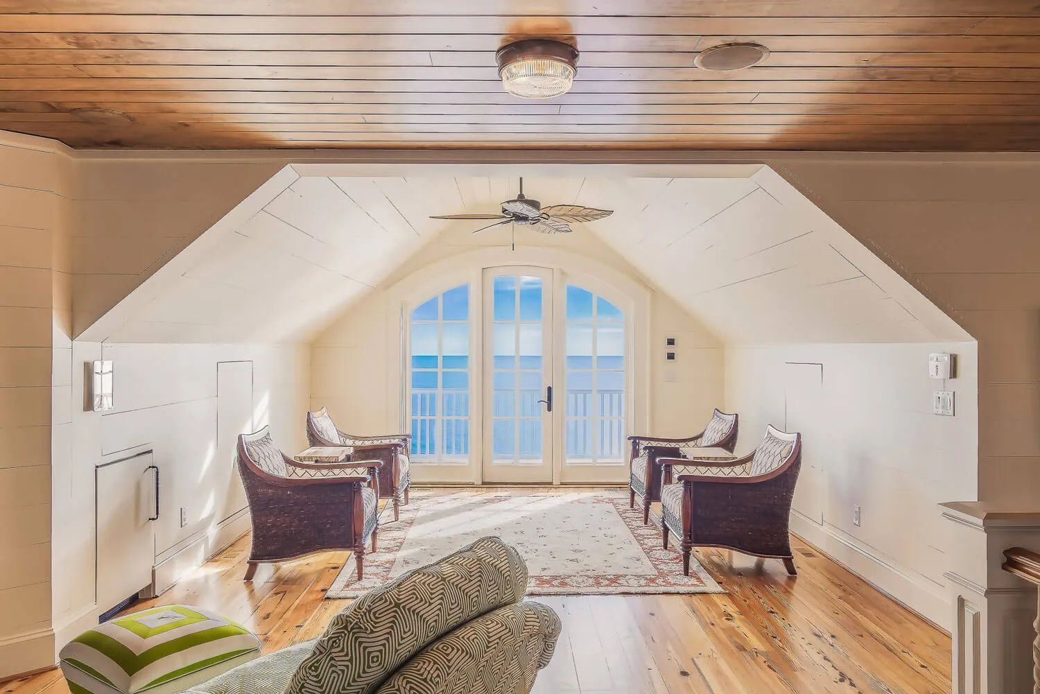 A sunlit attic room with a wooden ceiling, wicker chairs, an area rug, and doors leading to a balcony.