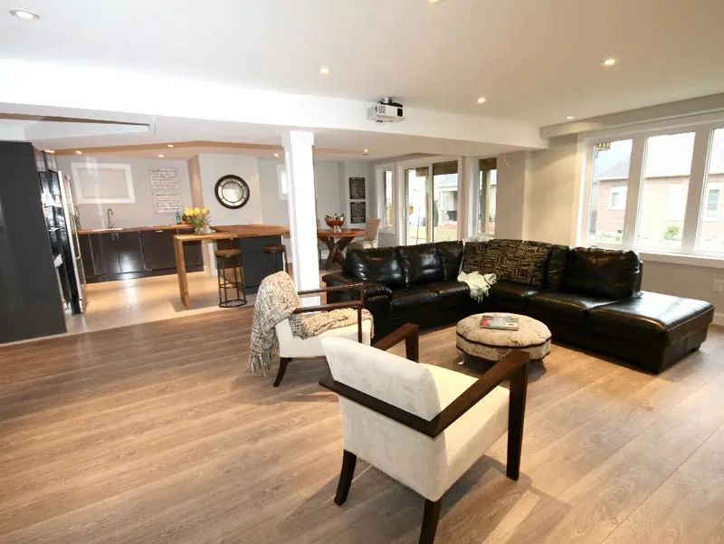 A modern living room with a black leather sectional, two light chairs, an ottoman, and a kitchen in the background.